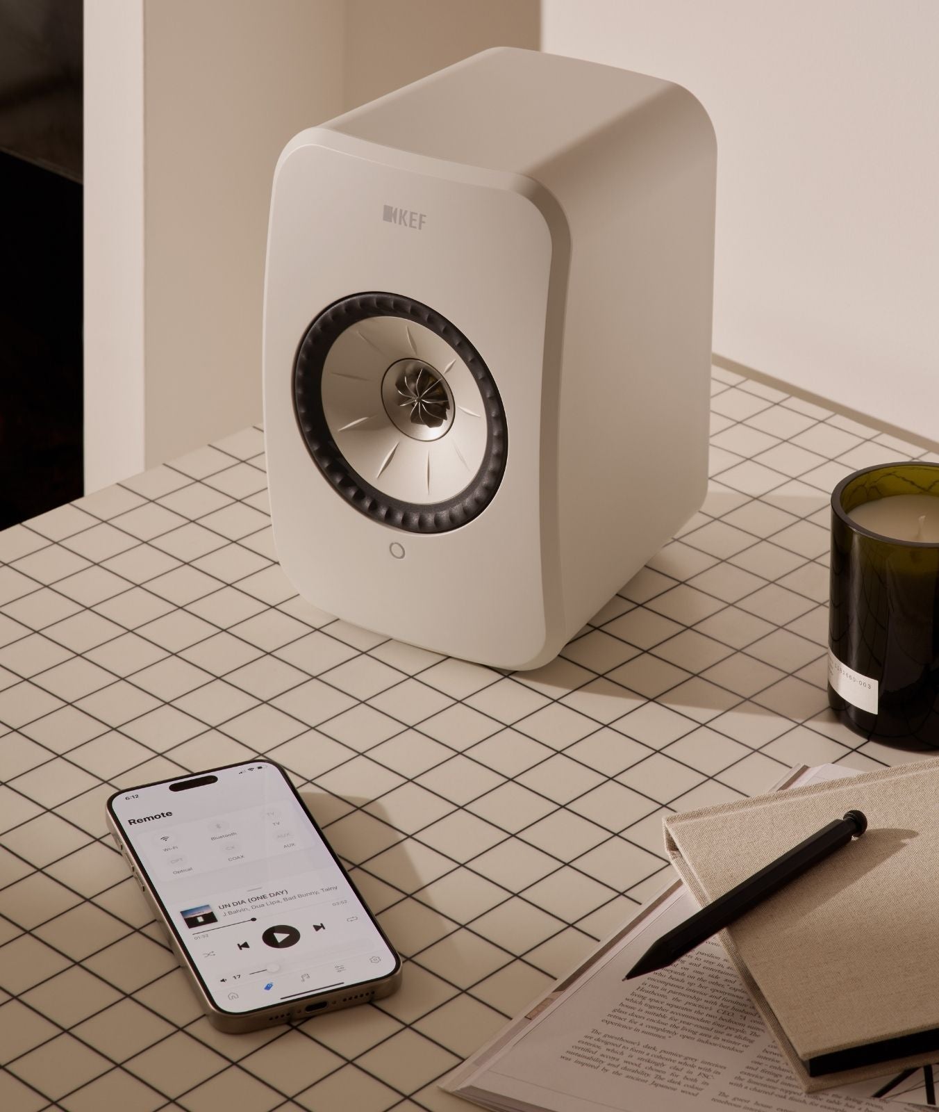 White LSX II LT speaker on a tiled desk with a phone, notebook, and candle nearby.