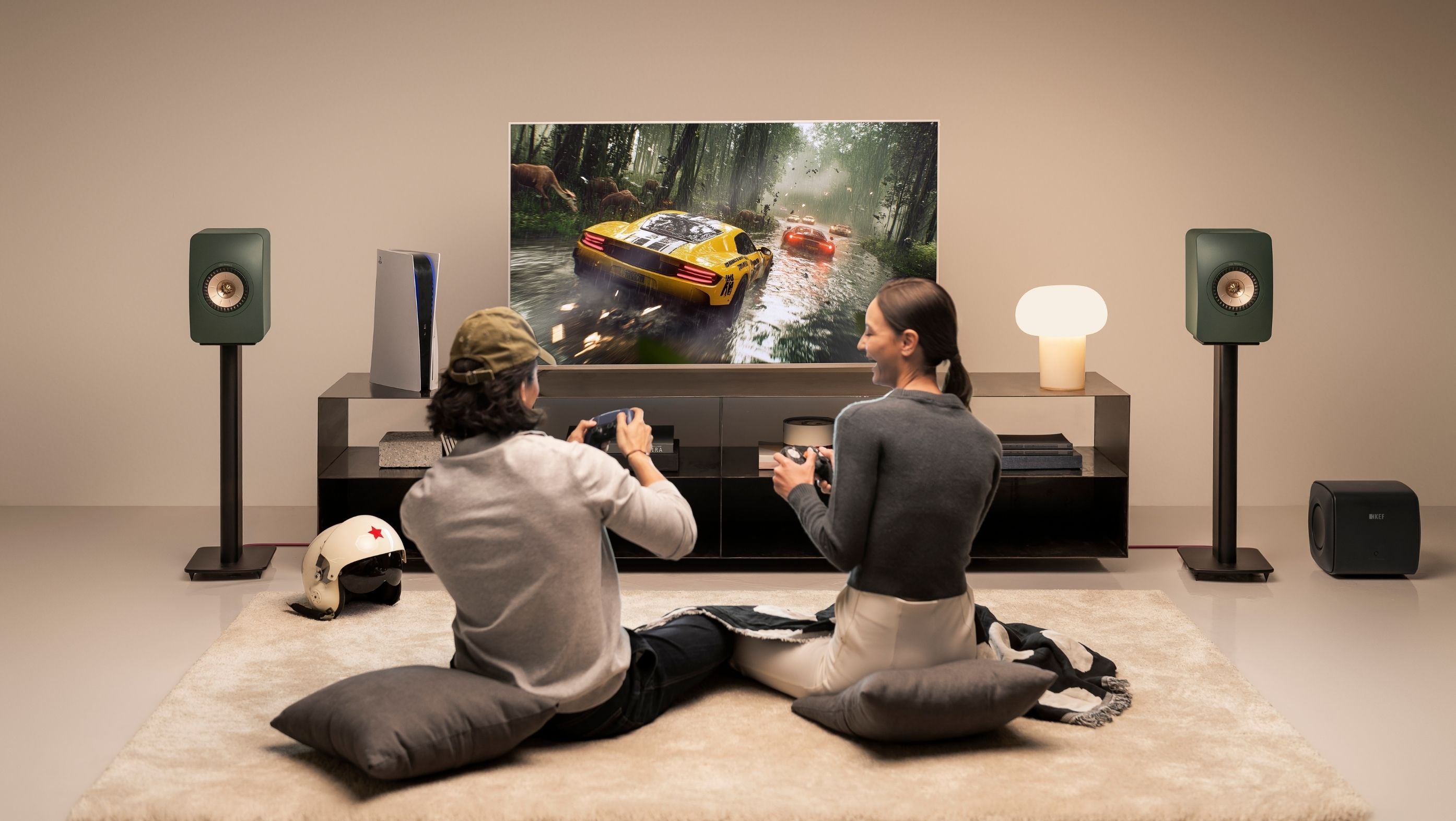 Two people sitting on the floor in front of a large TV and green LS50 Wireless II speakers playing a video game.